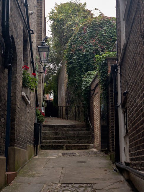 A narrow, uphill alleyway between brick buildings, featuring weathered stone steps leading to a higher level, surrounded by brick walls with black iron railings and outdoor lighting fixtures. The path